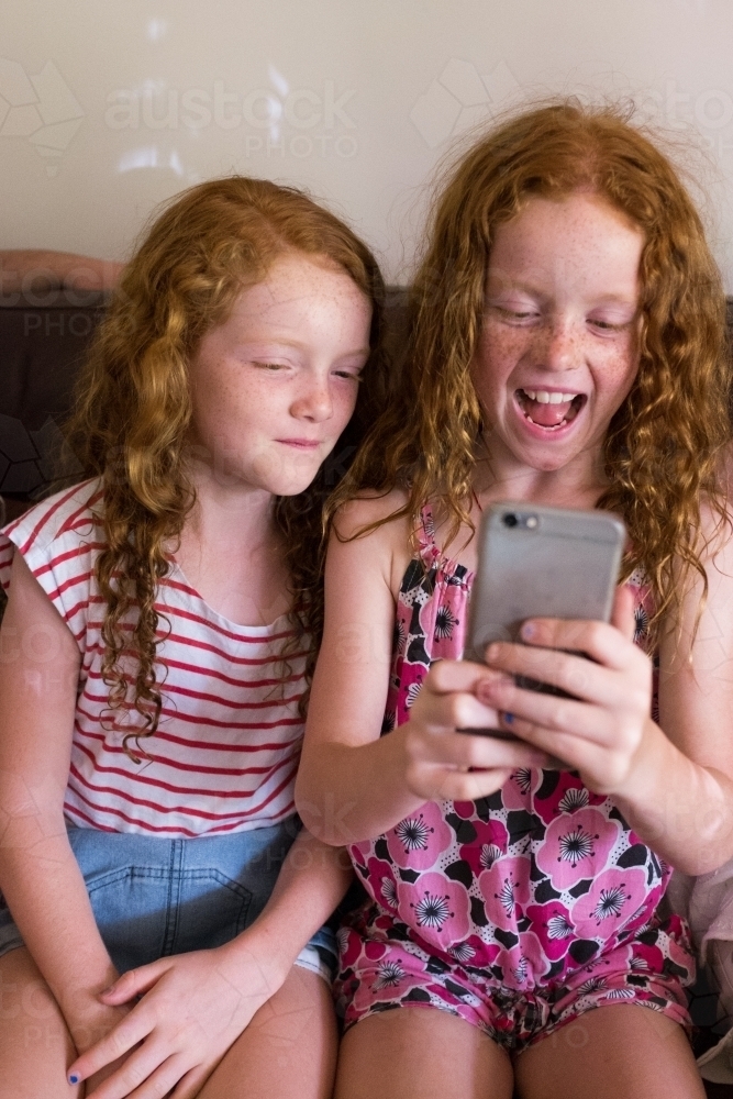Two girls sitting on a couch playing on a smartphone - Australian Stock Image