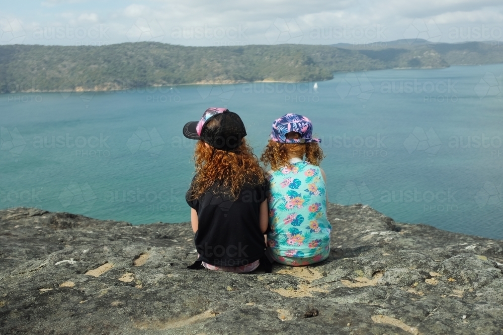 Image of Two girls sitting on a rock overlooking water - Austockphoto