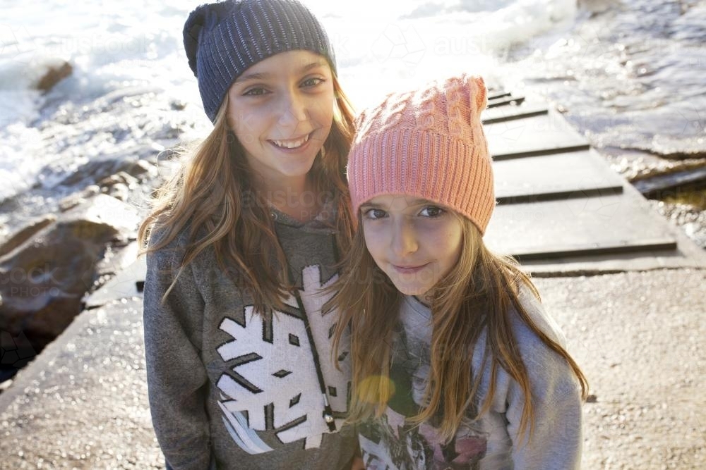 Two girls standing by the ocean looking up in the camera smiling - Australian Stock Image