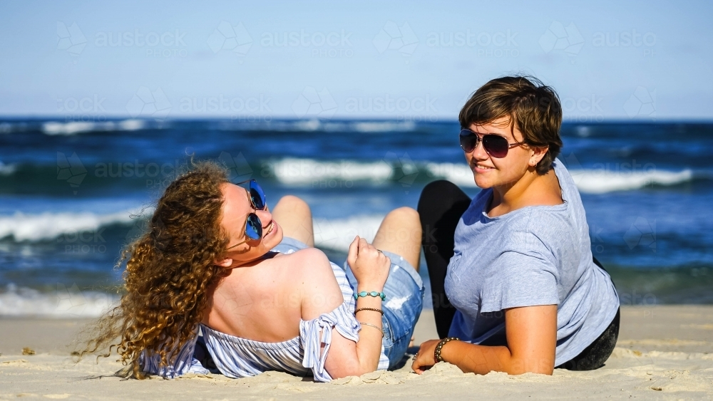 Two girls wearing sunglasses sitting on the sand - Australian Stock Image