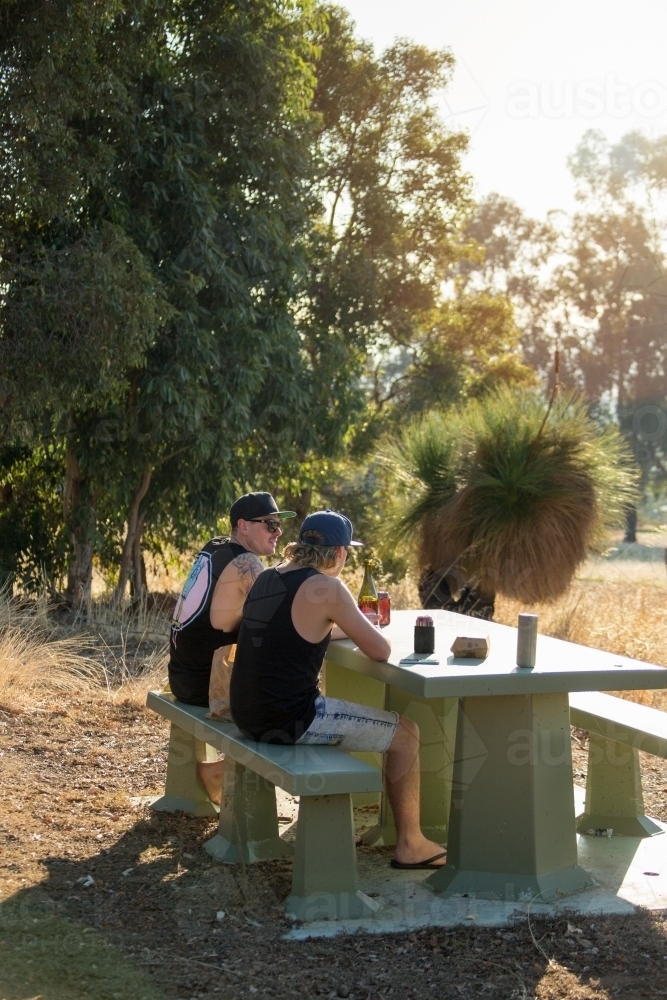 Two guys sitting at a picnic table with gum trees and grass trees in background - Australian Stock Image