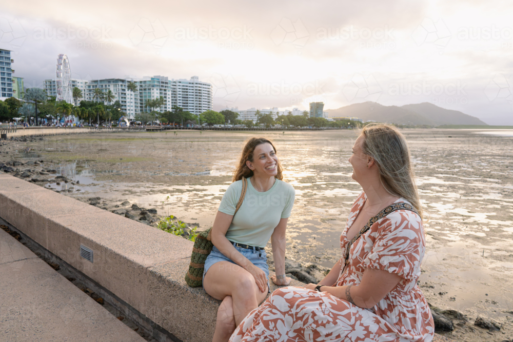 Two happy women talking side by side on a stone ledge overlooking the coastal landscape at sunset - Australian Stock Image