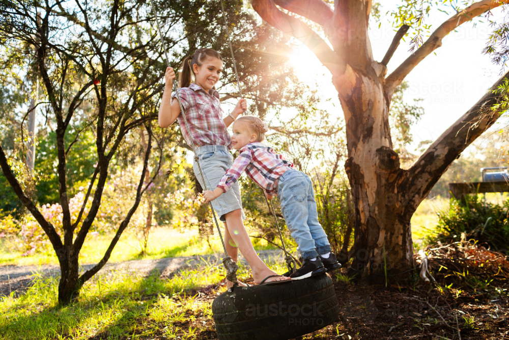 Two happy young Australian children swinging together in front yard of rural home on tyre swing - Australian Stock Image