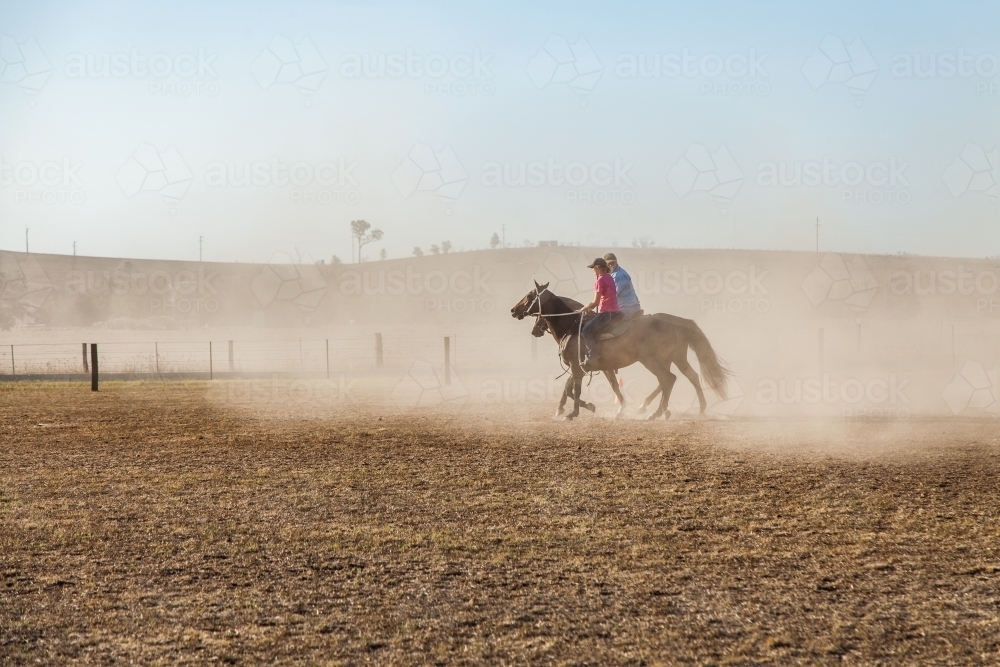 Two horses and riders riding through dusty paddock in summer - Australian Stock Image