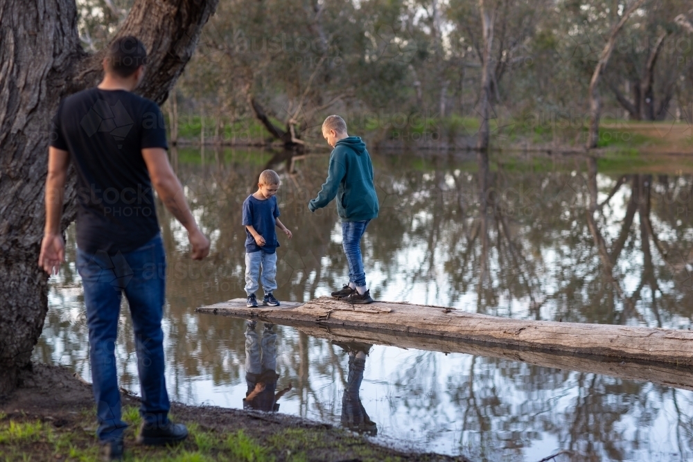 two kids balancing on log in water with father looking on in the foreground - Australian Stock Image