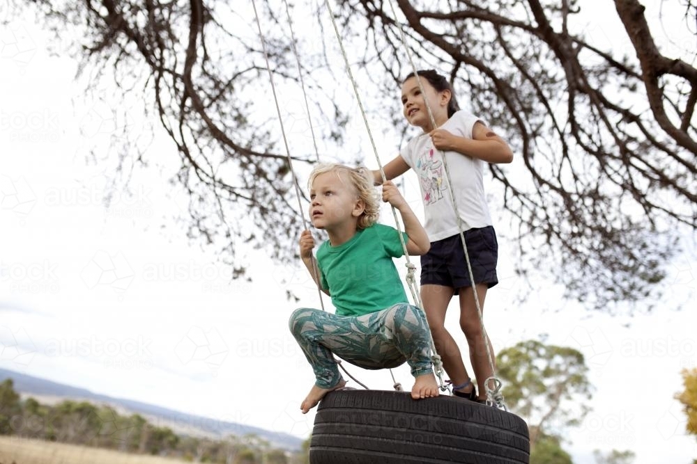 Two kids swinging on tyre swing in country back yard - Australian Stock Image