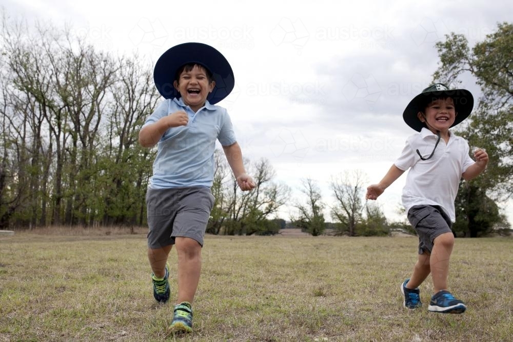 Two laughing boys wearing school uniform running across grass - Australian Stock Image