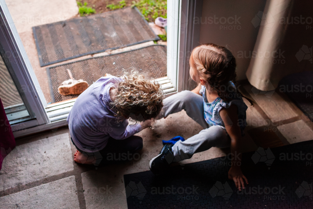 two little girls by back door of home helping each other get shoes on - Australian Stock Image