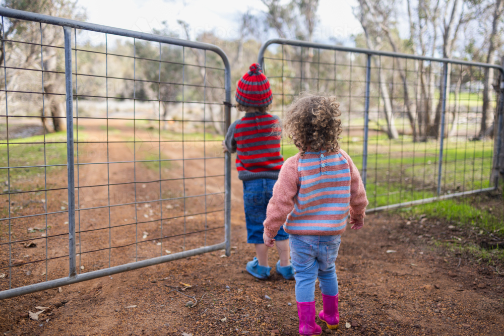 two little kids wearing warm woollen clothing standing at a closed gate on a rural property - Australian Stock Image