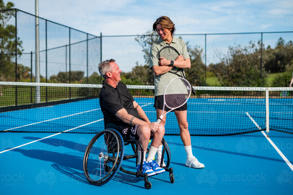 Two male tennis players, one in a wheelchair, laugh and engage on a blue court outdoors - Australian Stock Image