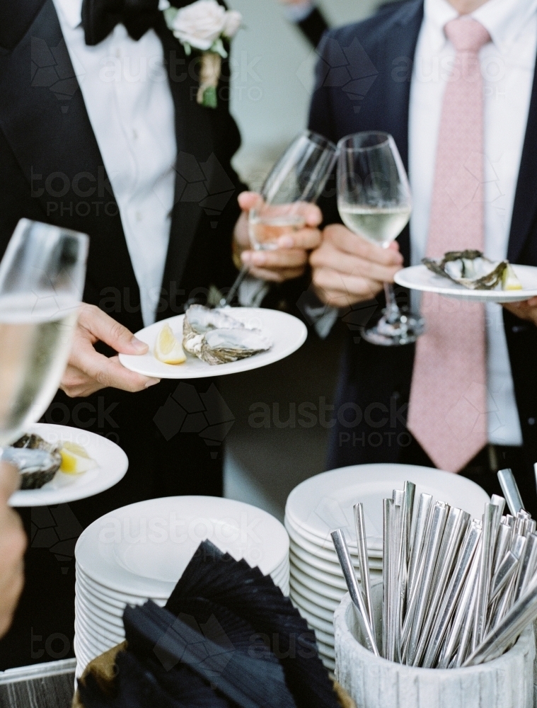 Two men in suits holding glasses of wine and plates of oysters at a wedding - Australian Stock Image