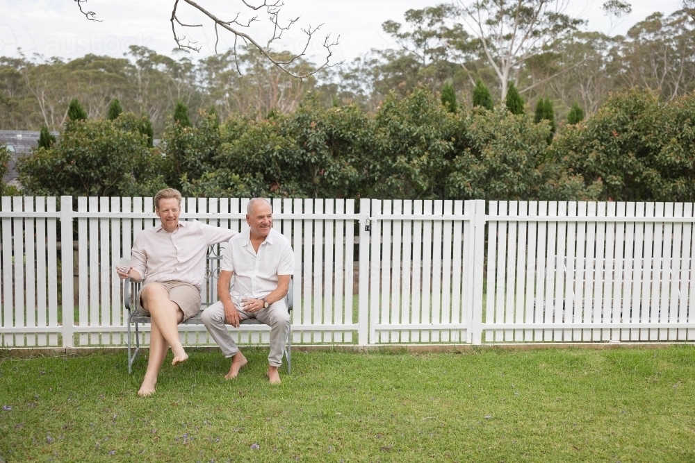 Two men sitting on backyard bench against white fence - Australian Stock Image