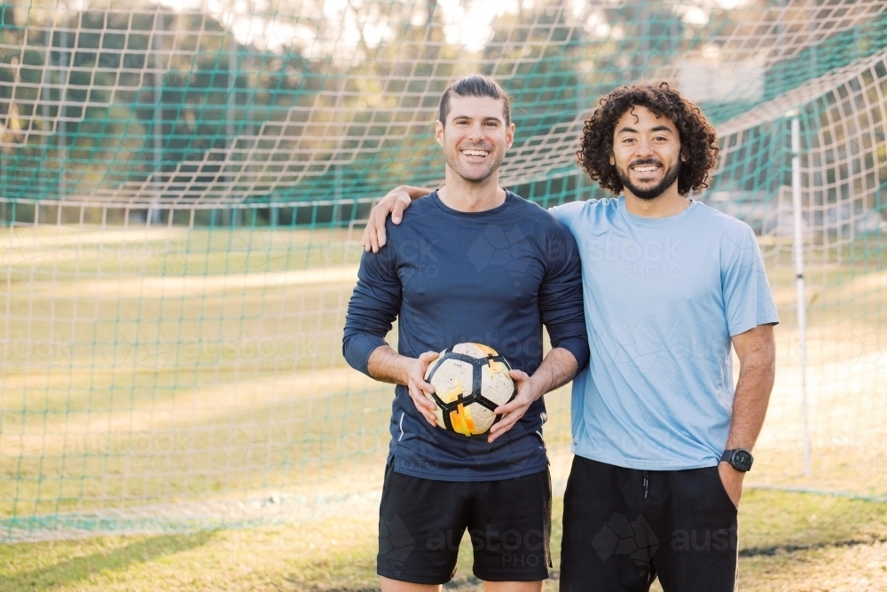 Two men smiling as they stand together in a soccer  goal, with one of the men holding a soccer ball - Australian Stock Image