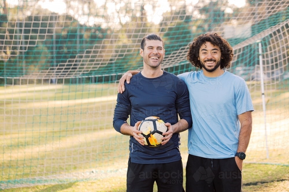 Two men smiling as they stand together in a soccer  goal, with one of the men holding a soccer ball - Australian Stock Image