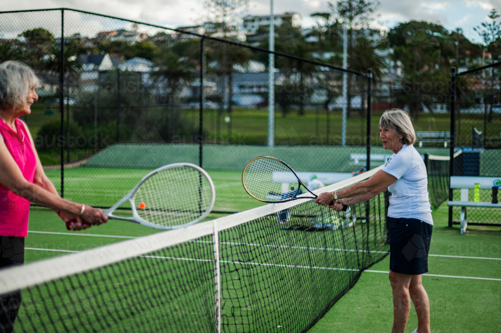 Two older women engage in a friendly tennis match at an outdoor court during the day - Australian Stock Image