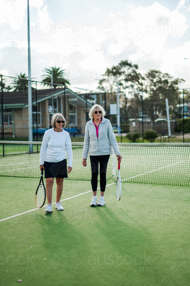 Two older women holding tennis racquets stand on a bright outdoor court in Australia. - Australian Stock Image