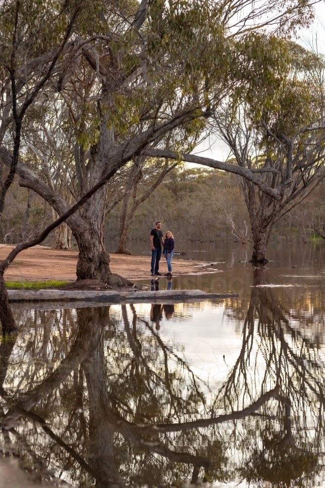 two people on edge of lake with gum trees reflected in water - Australian Stock Image