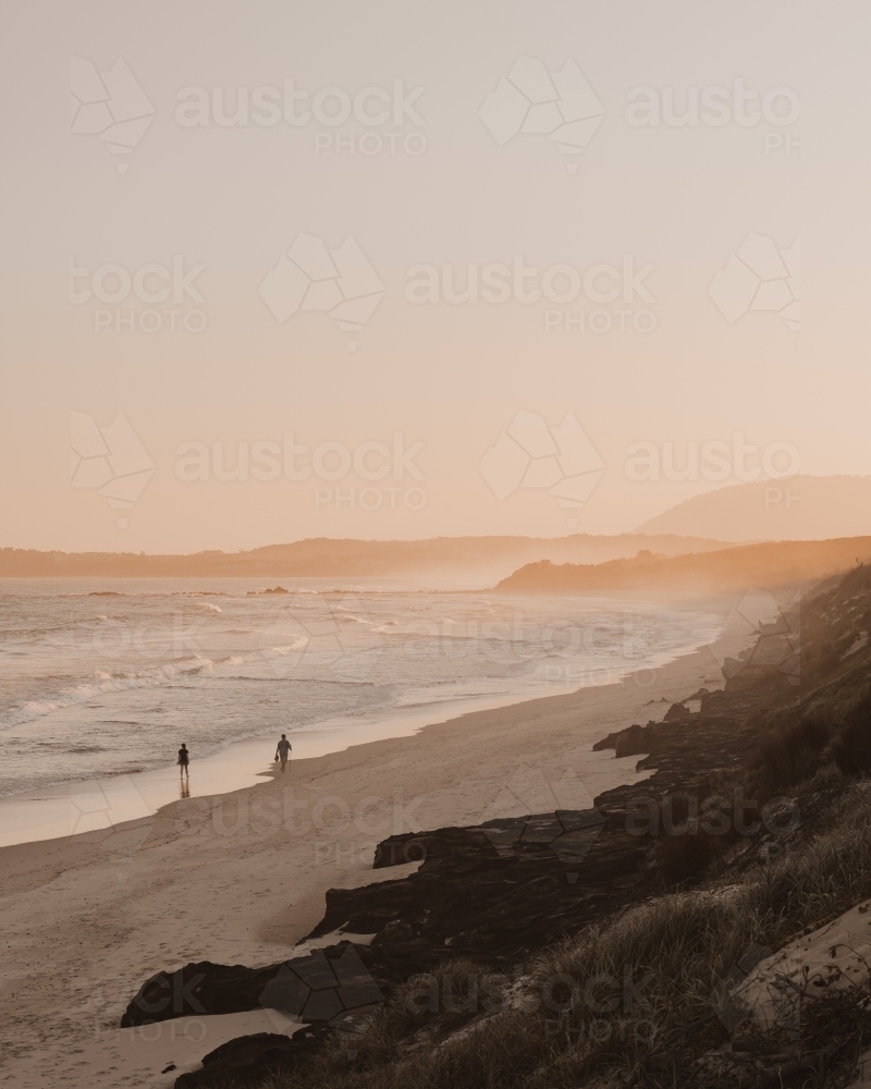 Two people walking along a deserted beach at sunset. - Australian Stock Image