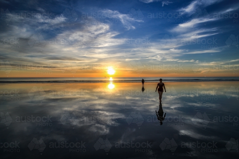 Two people walking along beach with sky reflected in water - Australian Stock Image