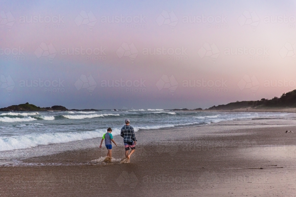 Two people walking in water on sand at beach at sunset from behind - Australian Stock Image