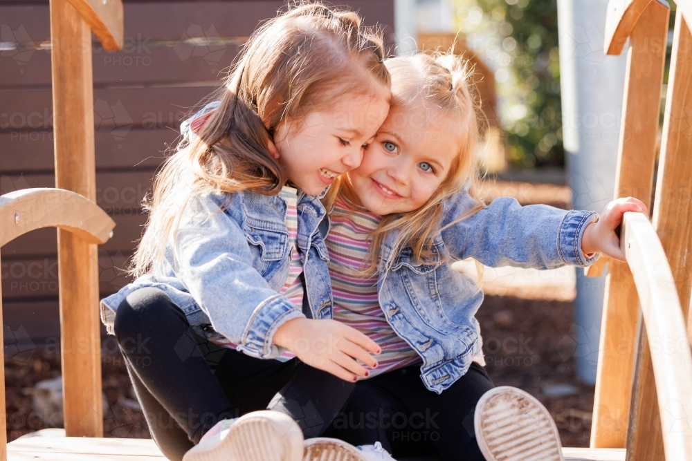 Two preschooler sisters sitting together on wooden bridge in kindergarten yard - Australian Stock Image