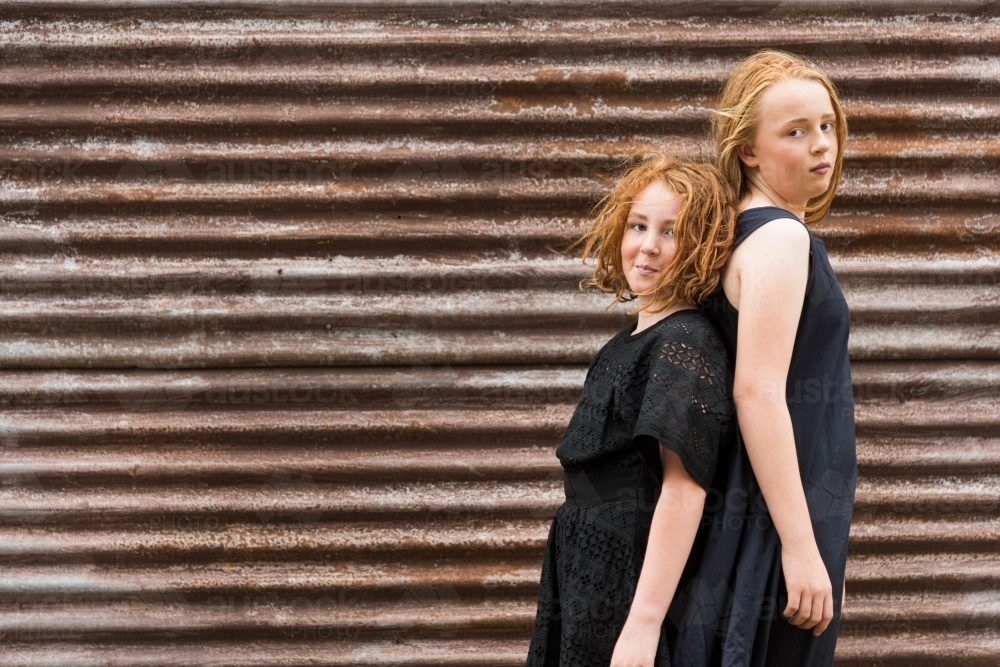 two red headed girls in black dresses back to back against rusty brown corrugated iron shed - Australian Stock Image