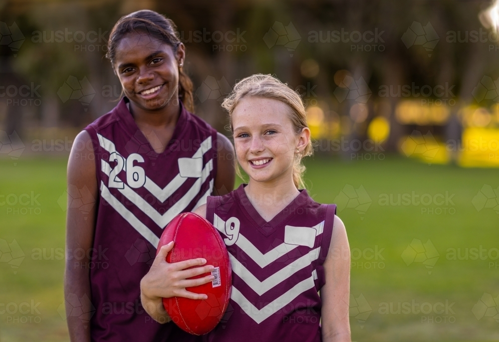 two school-aged girls in football jerseys holding aussie rules football - Australian Stock Image