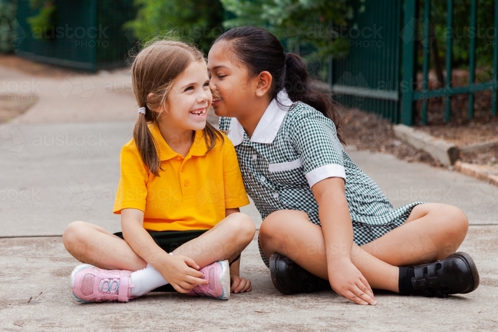 Two school girls sitting outside one child whispering secrets to other kid - Australian Stock Image