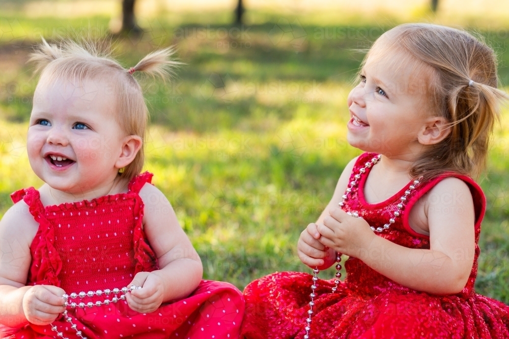 Two similar aged little girls in red Christmas dresses playing with strings of beads in garden - Australian Stock Image
