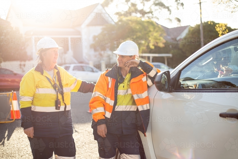 Two smiling women road workers with white helmet leaning on a white car - Australian Stock Image