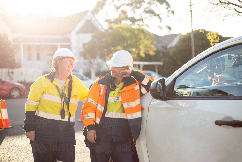 Two smiling women road workers with white helmet leaning on a white car - Australian Stock Image