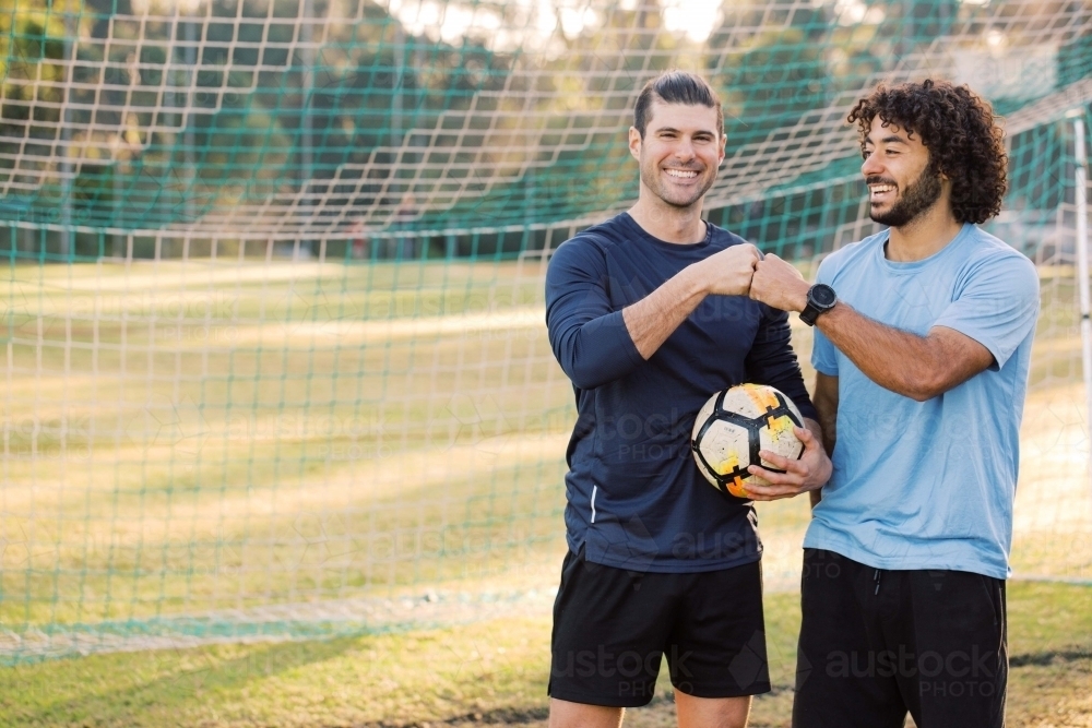 Two smiling young men standing on the field bumping their fists near the soccer net - Australian Stock Image
