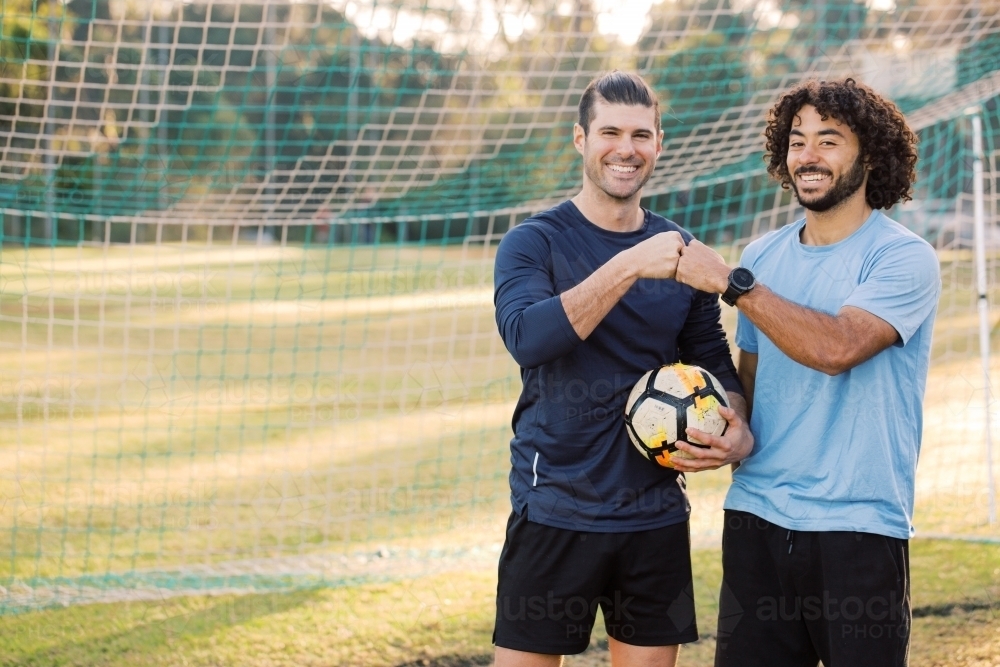 Two smiling young men standing on the field bumping their fists near the soccer net - Australian Stock Image