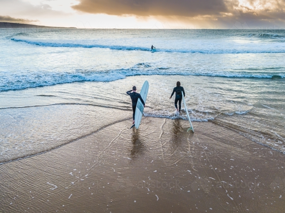 Two surfers walking into the ocean at sunrise - Australian Stock Image
