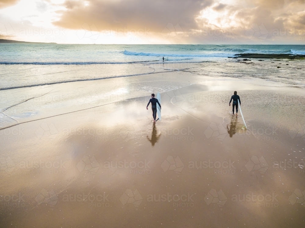 Two surfers walking on a wet beach into the ocean at sunrise - Australian Stock Image