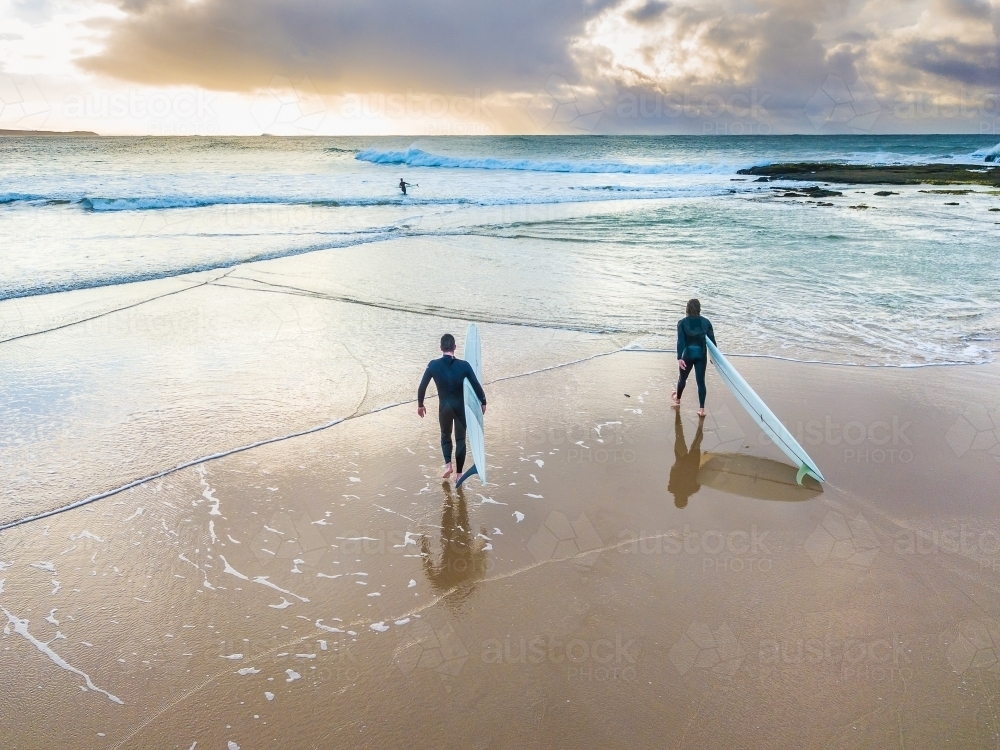 Two surfers walking on a wet beach into the ocean at sunrise - Australian Stock Image