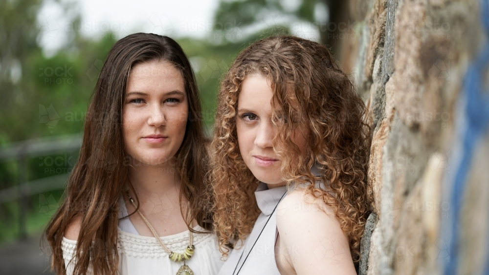 Two teen girls leaning against stone wall outdoors - Australian Stock Image