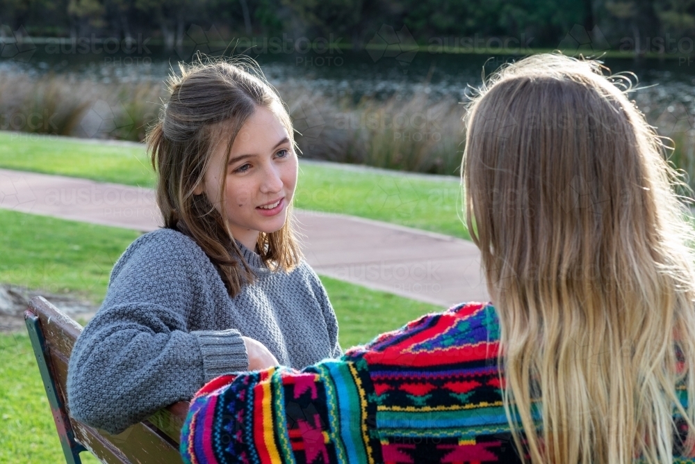 two teen girls sitting on park bench together - Australian Stock Image