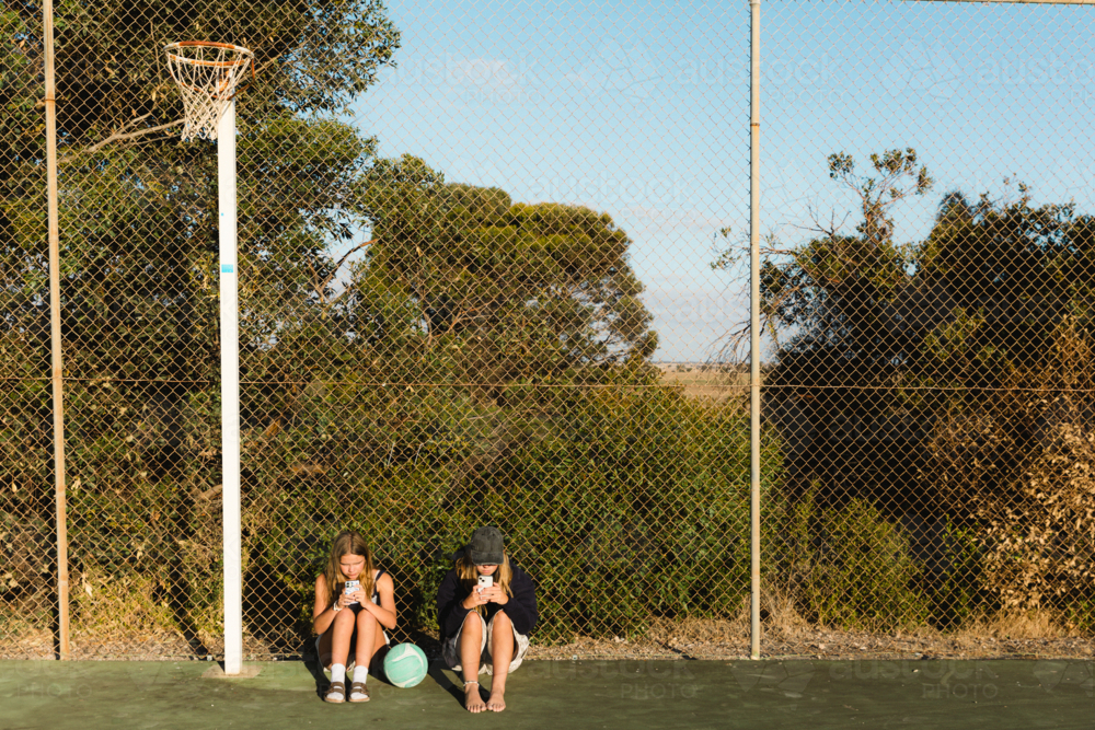 Two teenage girls on smart phones at regional outdoor netball court with ball - Australian Stock Image