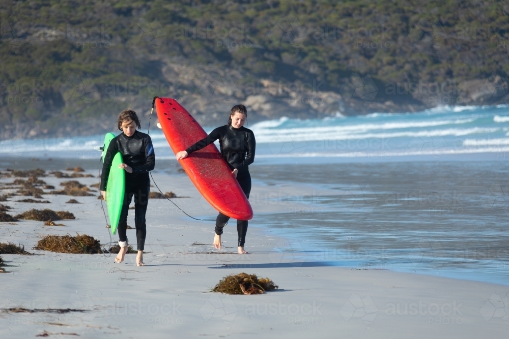 two teenagers in wetsuits carrying surfboards on the beach - Australian Stock Image