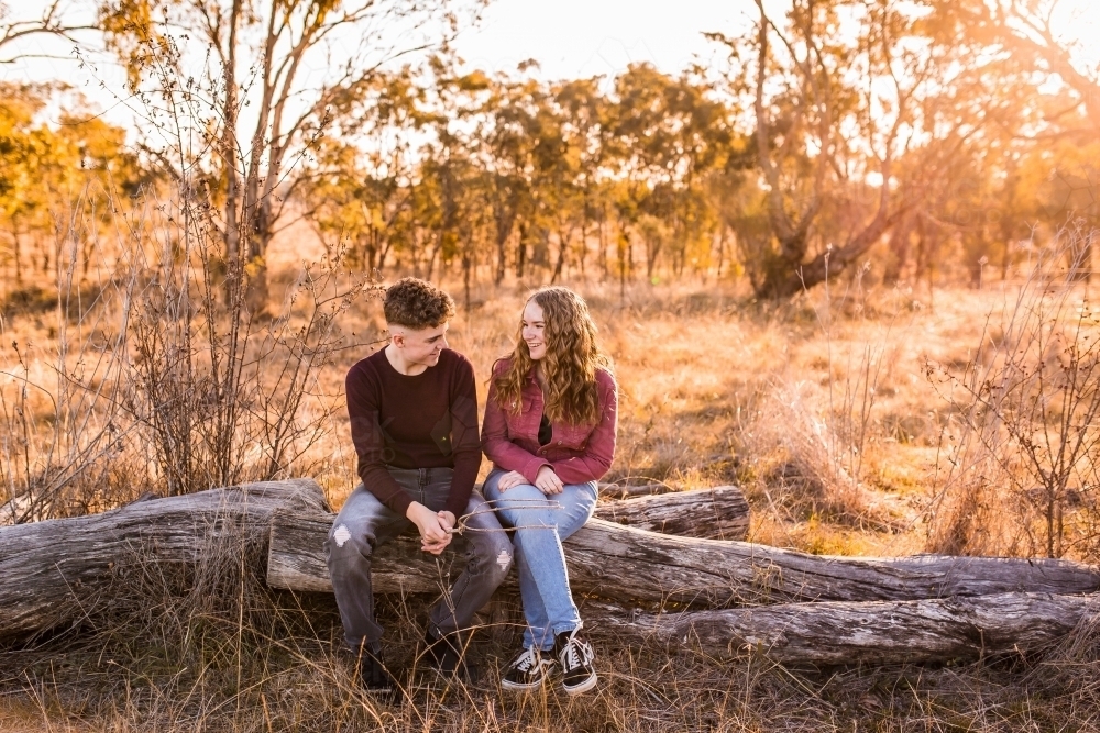 Two teenagers sitting on log in paddock talking and smiling - Australian Stock Image