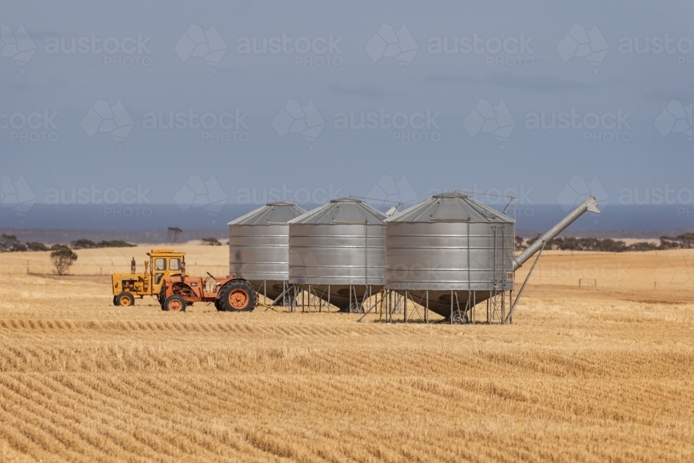 Two tractors & three metal grain silos in a patterned, harvested field - Australian Stock Image