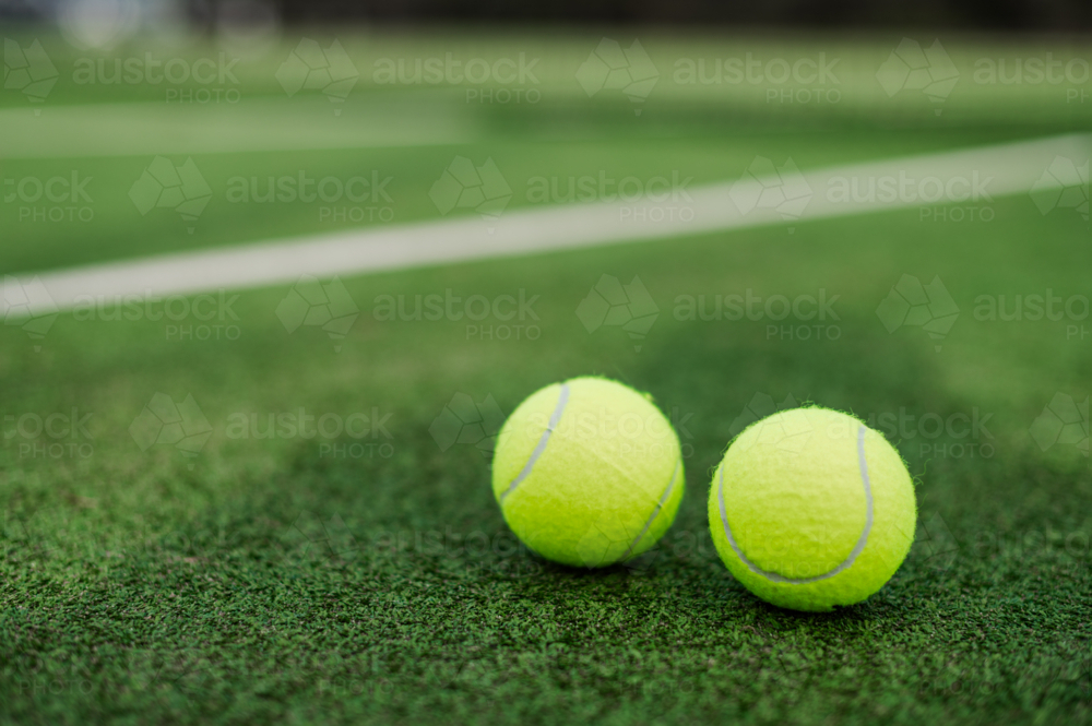 Two vibrant yellow tennis balls resting on a lush green tennis court in daylight - Australian Stock Image