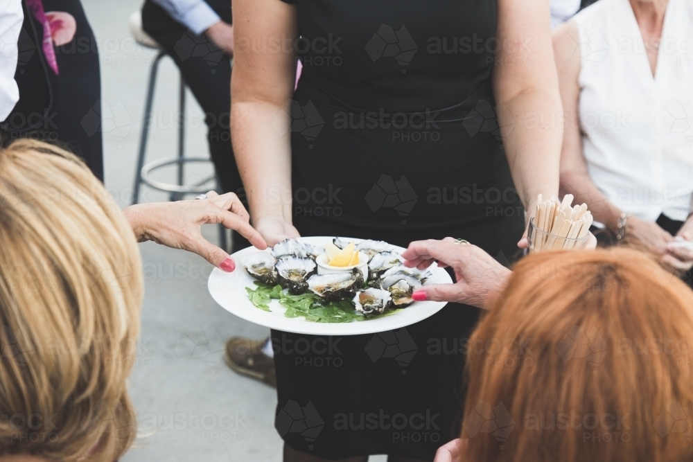 Two women's hands reaching for oysters from a platter being served by a waitress - Australian Stock Image