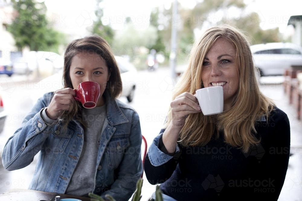 Two women sitting at a cafe drinking from their coffee cups and smiling - Australian Stock Image