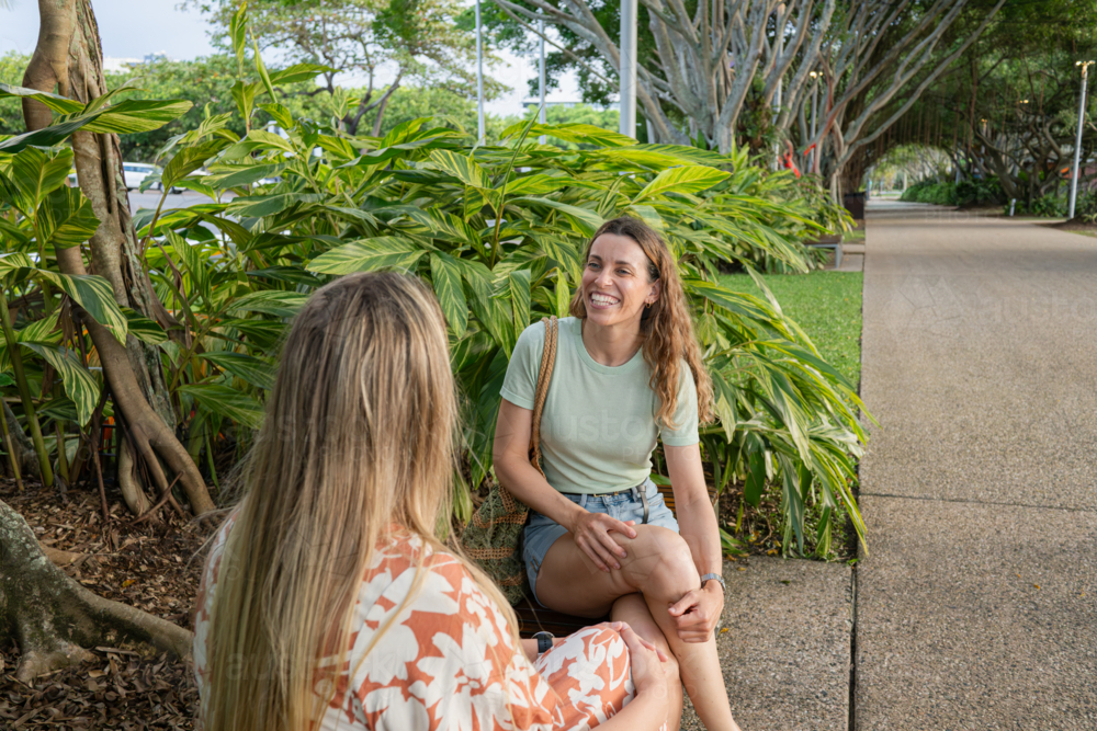 Two women sitting side by side along a tree-lined pathway in a lush park - Australian Stock Image