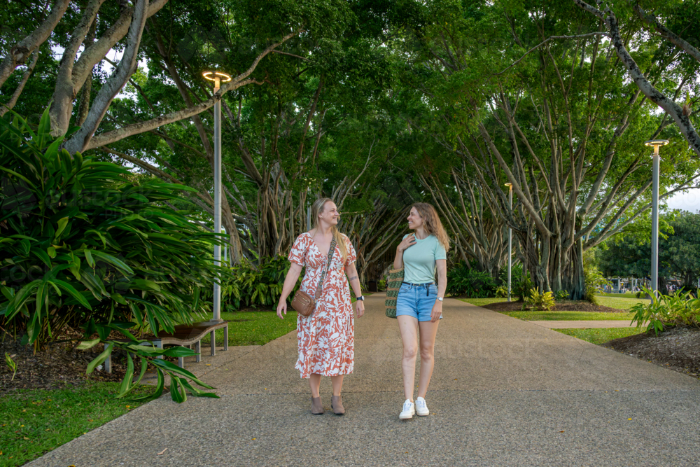 Two women walking side by side along a tree-lined pathway in a lush park - Australian Stock Image
