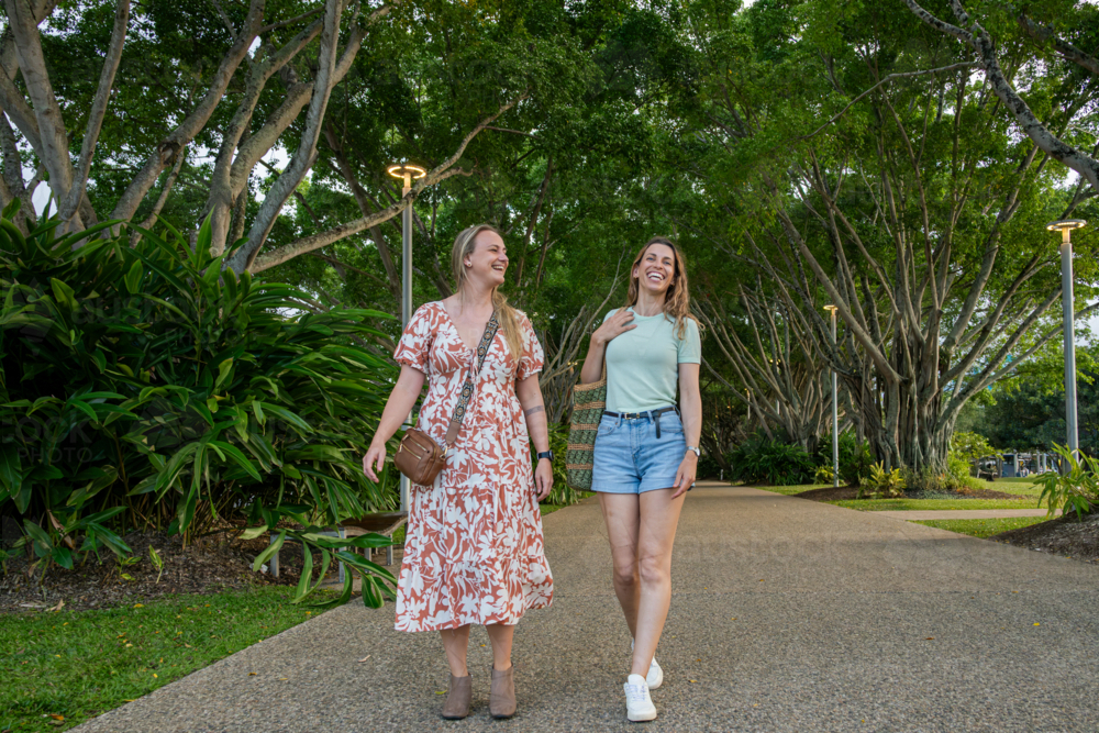 Two women walking side by side along a tree-lined pathway in a lush park - Australian Stock Image