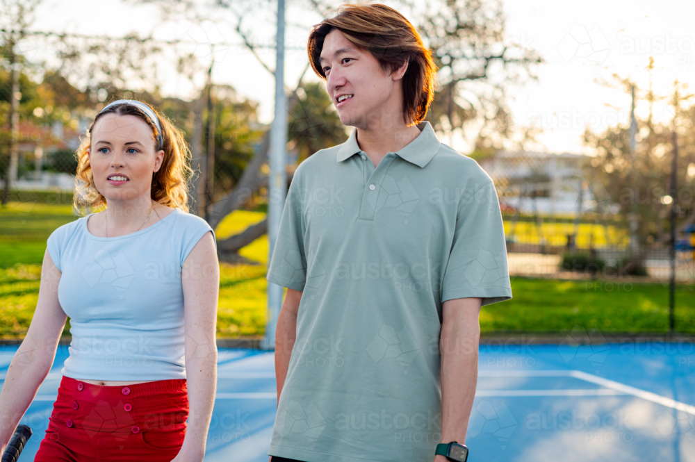 Two young adults chat on a tennis court, with green trees and a setting sun in the background - Australian Stock Image