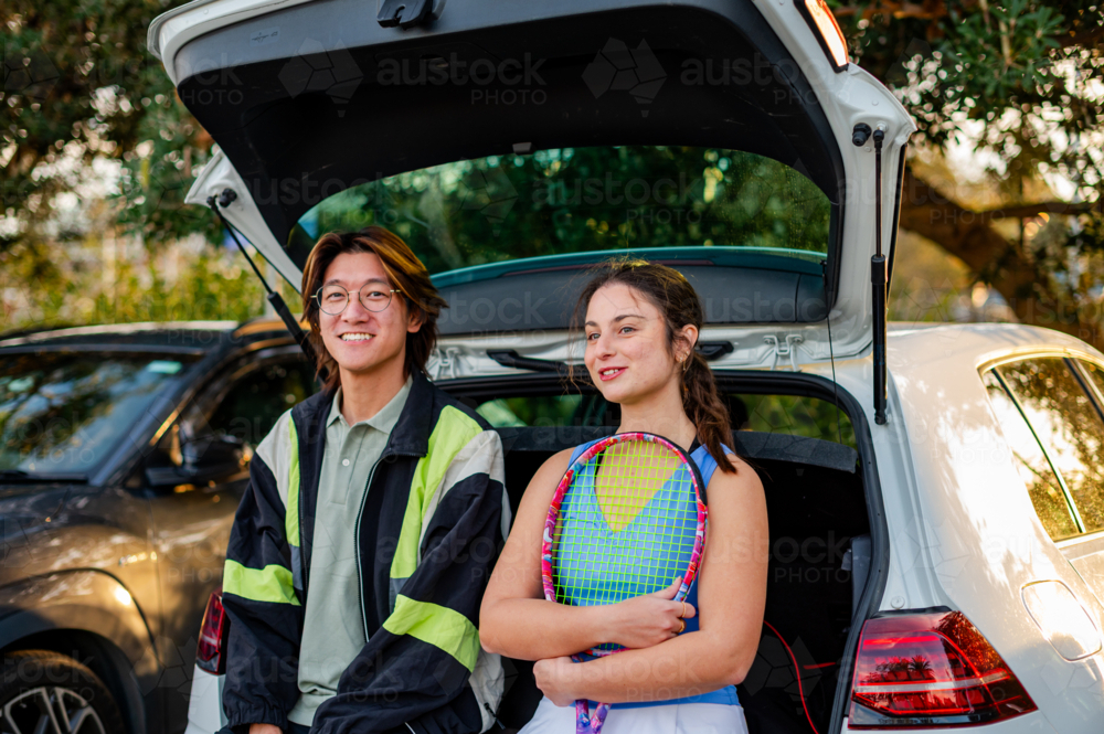 Two young adults relax by a car boot, ready to play tennis in a sunny park setting - Australian Stock Image